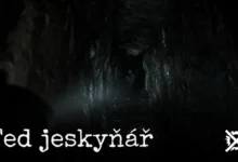 Temná úzká jeskynní chodba s průrvou ve skále a slabým světlem čelovky, v hloubi tmy náznak bílé postavy
