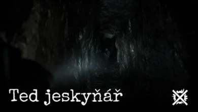 Temná úzká jeskynní chodba s průrvou ve skále a slabým světlem čelovky, v hloubi tmy náznak bílé postavy